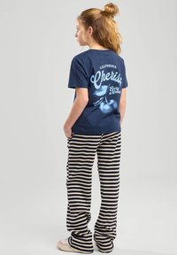 T-shirt di cotone blu navy con grafica e testo sul retro; abbinato a pantaloni rigati marroni e crema. Calzature casual visibili.