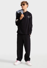 Zwarte langemouwen shirt met een grafisch logo, gecombineerd met gestructureerde zwarte broek. Witte sneakers met roze veters en een spijkerjack over één schouder gehangen.