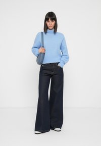 Svetlo modrý pletený sveter s vysokým golierom, v kombinácii s tmavými džínovými nohavicami so širokými nohavicami. Outfit dopĺňa malá modrá crossbody taška.