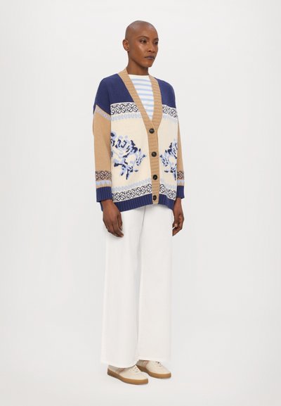 Strickcardigan in Beige, Marineblau und Hellblau mit floralem Muster, Knopfverschluss und gerippten Bündchen, kombiniert mit weißen weit geschnittenen Hosen und Sneakers.