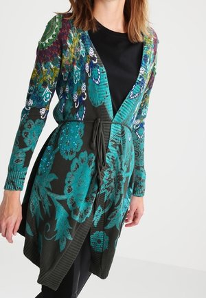 Femme portant un long cardigan en tricot avec des motifs floraux turquoise et une taille à cordon sur une chemise et un pantalon noirs.