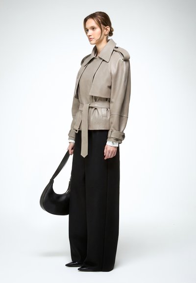 Nő bézs színű, övvel ellátott bőrkabátban és fekete, bőszárú nadrágban, kezében fekete kézitáskával, egyszerű fehér háttér előtt áll.