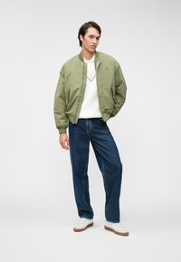 Πράσινο bomber μπουφάν με ριμπ κολάρο, λευκή ζακέτα με σχέδιο V, μπλε τζιν παντελόνι και άσπρα αθλητικά παπούτσια με ανοιχτό καφέ σόλες.