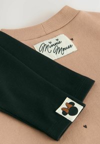 Beige topp med ribbad halsringning märkt "Minnie Mouse" och svarta ribbade ärmar som visar en silhuett av Minnie Mouse med en brun rosett.