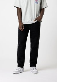 Camiseta de algodón gris para hombre con mangas cortas y logo azul, combinada con jeans de mezclilla negros y zapatillas blancas. Corte plano y casual.