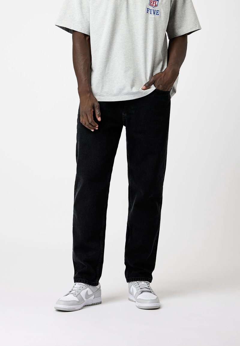 Camiseta de algodón gris para hombre con mangas cortas y logo azul, combinada con jeans de mezclilla negros y zapatillas blancas. Corte plano y casual.