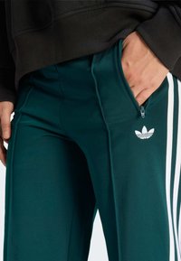 Teal athletic pants feature white side stripes, zippered pocket, and an embroidered white logo. The fabric is smooth with a slight sheen.