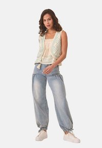 Chaleco ligero de tejido con patrones geométricos en menta sobre una base crema, combinado con pantalones de denim azul claro holgados y zapatillas blancas.