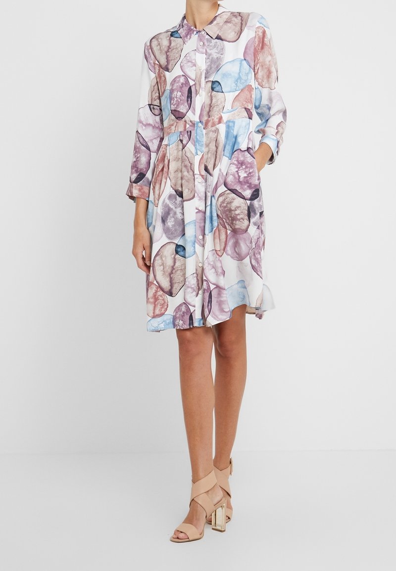 Femme portant une robe mi-longue boutonnée à manches longues avec un motif abstrait multicolore, associée à des sandales beige à talons hauts.