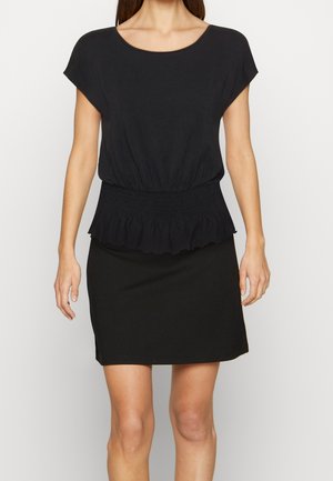 Femme vêtue d'un haut noir à manches courtes avec une taille froncée et d'une jupe noire assortie longueur genou, debout devant un fond uni.