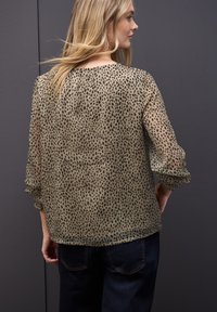 Vrouw met lang blond haar, gekleed in een beige blouse met lange mouwen en stippen, en een donkere spijkerbroek, staand tegen een donkergrijze achtergrond.