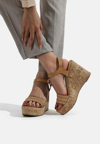 Sandali con zeppa e piattaforma in sughero, con una parte superiore marrone e fasce intrecciate. Presentano un dettaglio decorativo e una cinghia alla caviglia con chiusura a fibbia.