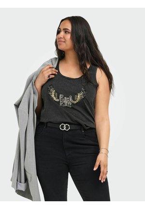 Kvinde med langt mørkt hår iført en sort "Rock & Roll" tanktop og sorte bukser, der holder en lysegrå blazer over den ene skulder.