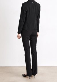 Black fitted blazer and matching trousers, smooth fabric, structured design, with slits at the trouser hems, paired with black heels.