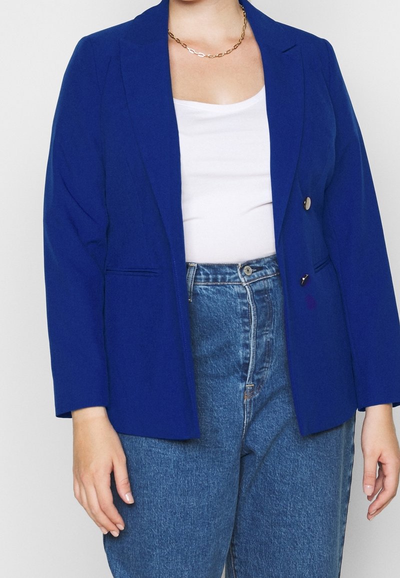 Blazer azul hecho de tela texturizada, con cierre de un solo botón, solapas de muesca y dos bolsillos frontales. Combinado con una blusa blanca y jeans.