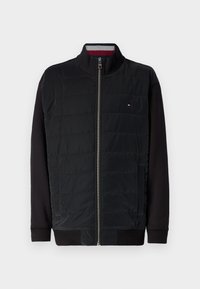 Black jacket with a quilted front and sweater-like sleeves. Features a high collar, zip closure, and small logo on the chest.