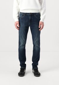Jeans de mezclilla azul oscuro con un corte clásico, que presentan un diseño de cinco bolsillos, un desgaste sutil y costuras en contraste. Llevados con zapatillas negras.