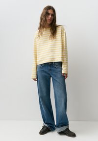 Femme aux cheveux longs et ondulés portant un chemisier à manches longues rayé jaune et blanc, un jean à jambes larges retroussé, et des chaussures bateau noires, debout contre un mur blanc.