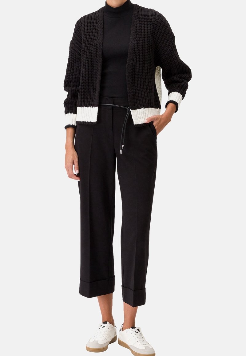 Svart stickad cardigan med vita manschetter, matchad med svarta vida byxor och vita sneakers. Outfiten har en enkel, avslappnad design.