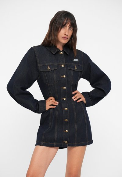 Δερμάτινο φόρεμα σε σκούρο μπλε με μακριά μανίκια, εφαρμοστή κοπή, μπροστινές κουμπώσεις, δύο τσέπες στο στήθος και χρυσές λεπτομέρειες.