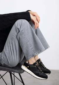 Las zapatillas negras y grises presentan una mezcla de materiales, incluyendo ante y malla, con acentos dorados y una suela blanca. Llevadas con pantalones de pierna ancha grises.