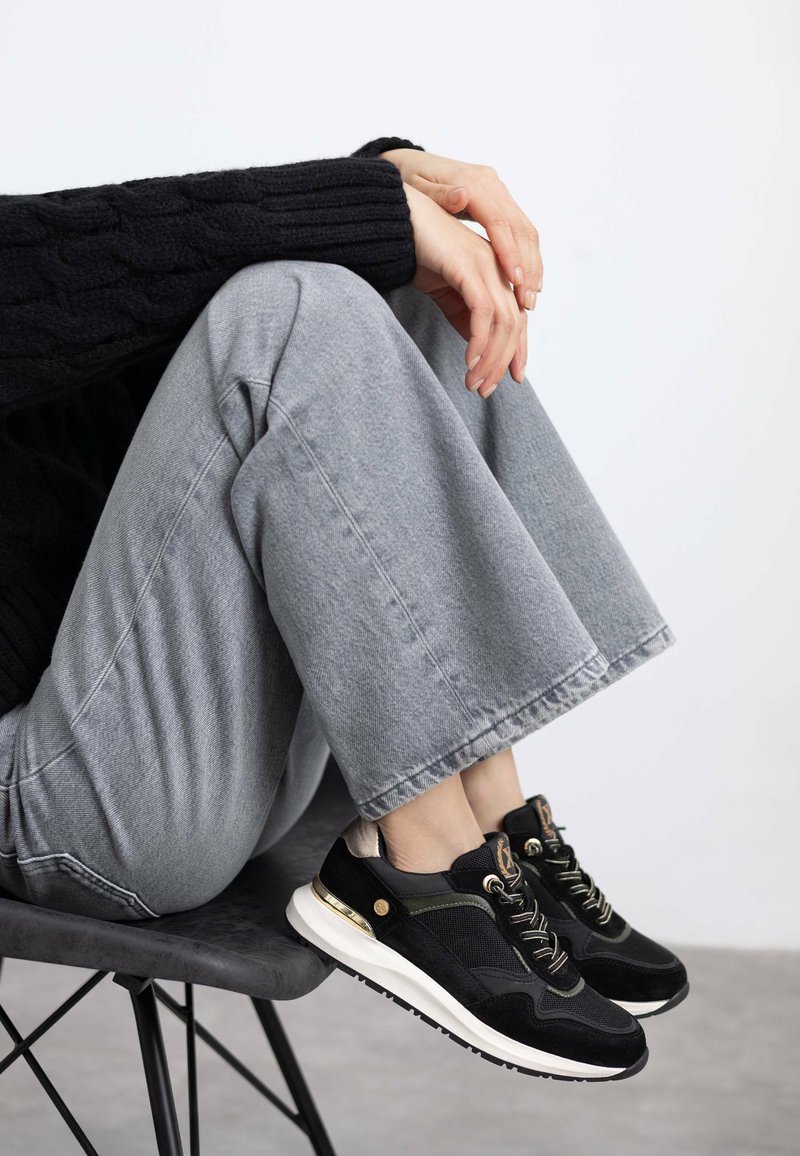 Las zapatillas negras y grises presentan una mezcla de materiales, incluyendo ante y malla, con acentos dorados y una suela blanca. Llevadas con pantalones de pierna ancha grises.