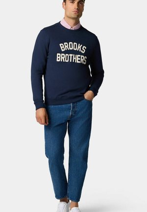 Felpa girocollo navy con la scritta "BROOKS BROTHERS" in lettering crema, abbinata a jeans in denim blu chiaro e sneakers bianche.