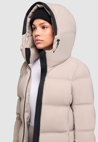 Chaqueta acolchada beige con una gran capucha, acentos negros y cierre de cremallera. Presenta una textura acolchada y un cordón ajustable en la capucha.