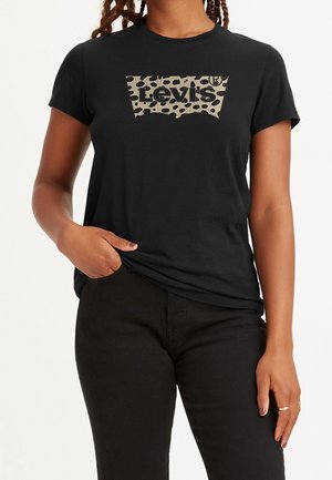Kobieta w czarnej koszulce Levi's z beżowo-czarnym logo w panterkę, zestawiona z czarnymi spodniami, stoi z jedną ręką w kieszeni.