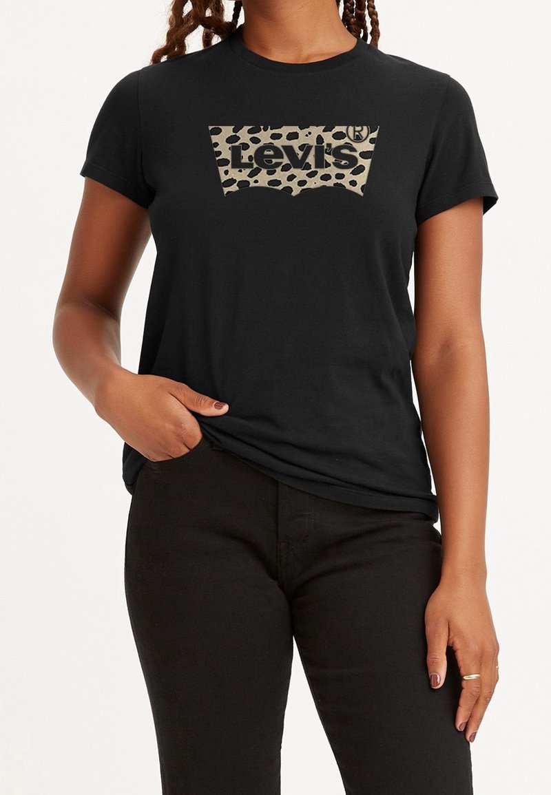 Femme portant un t-shirt noir Levi's avec un logo léopard beige et noir, associé à un pantalon noir, debout avec une main dans la poche.