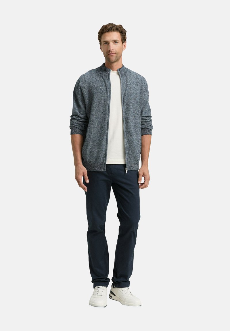 Graue strukturierte Bomberjacke, weißes T-Shirt, marineblaue Hose und weiße Sneakers. Die Jacke hat einen Reißverschluss und gerippte Bündchen.