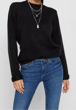 Personne portant un pull noir à manches longues, un jean bleu et des colliers chaînes argentés superposés sur un fond uni.