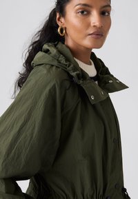 Veste à capuche vert olive en tissu léger avec cordons de serrage, boutons-pression et ourlet élastique. Présente une coupe décontractée et un design minimaliste.