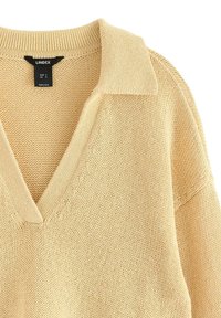 Kortærmet v-hals sweater i lys beige. Fremstillet af tykt strikket stof med ribkant i kraven og løs pasform. Har ingen yderligere mønstre eller detaljer.