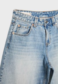 Jean en denim bleu clair avec un design à cinq poches, rivets en cuivre, passants de ceinture et une fermeture par bouton métallique, taille 36 visible sur l'étiquette intérieure.