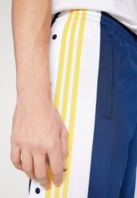 Navy och vita sportbyxor med gula ränder. Funktioner inkluderar ett elastiskt midjeband, sidofickor och ett strukturerat tyg.