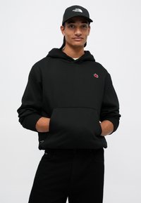 Mladý muž s copánky, který má na sobě černou mikinu s červeným logem a černou kšiltovku značky North Face, má ruce v kapse mikiny.
