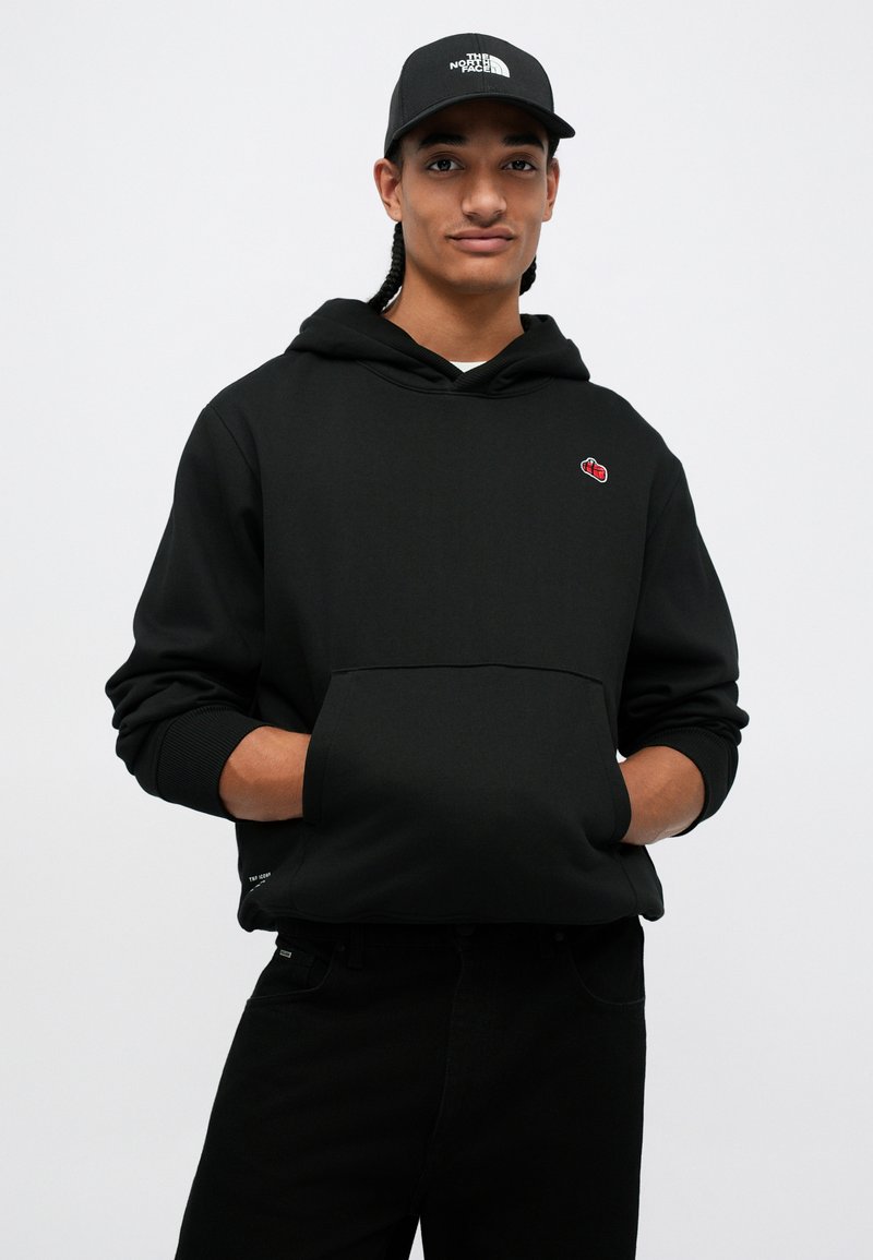 Jeune homme avec des cheveux tressés portant un sweat à capuche noir avec un logo rouge et une casquette noire The North Face, les mains dans la poche du sweat.