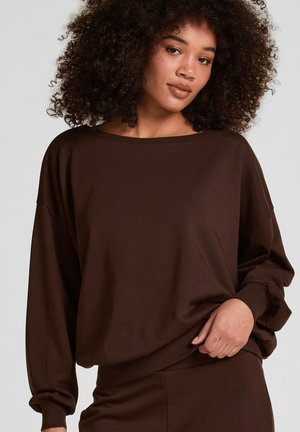 Vrouw met krullend haar die een losse donkerbruine top met lange mouwen en bijpassende broek draagt, terwijl ze de top iets bij de taille optrekt.