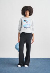 Mladá žena s kudrnatými vlasy, která má na sobě šedou mikinu s potiskem boty a nápisem „Kdo řekl, že žena nebyla stvořena k létání“, černé kalhoty, drží modrou čepici.