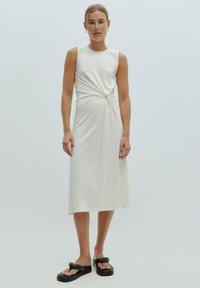 Witte, mouwloze jurk met een gedraaide knoopdetail op de taille. Glad stof, middellange lengte, gecombineerd met zwarte plateausandalen.