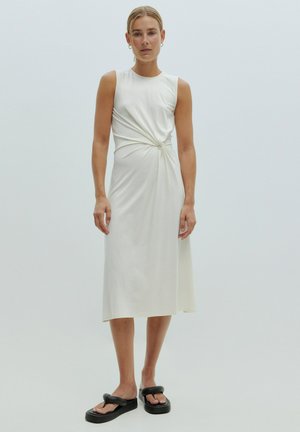 Witte, mouwloze jurk met een gedraaide knoopdetail op de taille. Glad stof, middellange lengte, gecombineerd met zwarte plateausandalen.