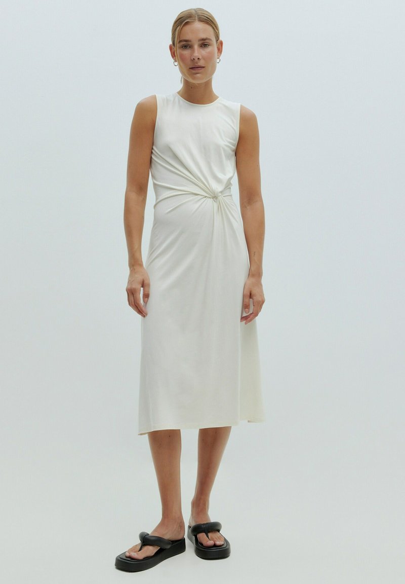 Witte, mouwloze jurk met een gedraaide knoopdetail op de taille. Glad stof, middellange lengte, gecombineerd met zwarte plateausandalen.