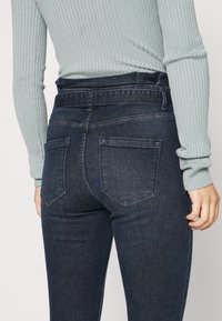 Donna che indossa jeans attillati a vita alta di colore blu scuro e un top a maniche lunghe ribattuto di colore blu chiaro, mostrata di schiena all'altezza della vita.