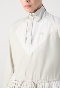 Vit lättviktsjacka med hög krage, dragkedjeöppning och veckad fåll; har dragsnören och broderad logotypdetalj.