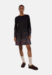 Abito nero con maniche lunghe, motivi floreali all'orlo e colletto rotondo. Abbinato a calze legwarmers lavorate a maglia di colore chiaro e pantofole marroni.