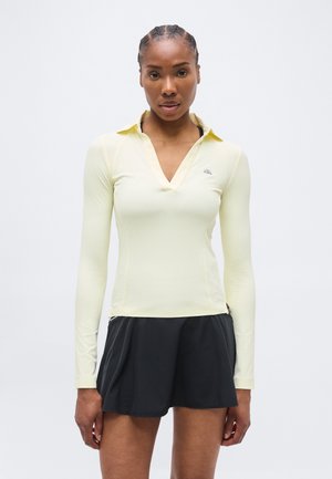 Femme aux cheveux tressés portant un haut à manches longues de couleur jaune pâle avec col et un short de sport noir, se tenant devant un fond clair uni.