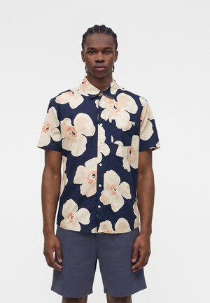 Νέος άνδρας με πλεξούδες φοράει μπλε πουκάμισο με μεγάλα μπεζ λουλούδια και σκούρα μπλε σορτς, στέκεται μπροστά σε μονόχρωμο φόντο.