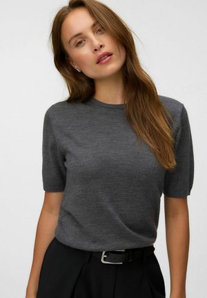 Femme aux longs cheveux bruns portant un pull à manches courtes gris foncé et un pantalon noir avec une ceinture noire, sur un fond uni.
