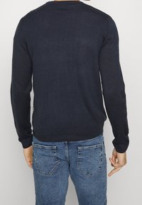 Donkerblauwe gebreide trui met lange mouwen en een ronde hals, met ribgebreide zoom en manchetten, gezien van de achterkant in combinatie met spijkerbroek.